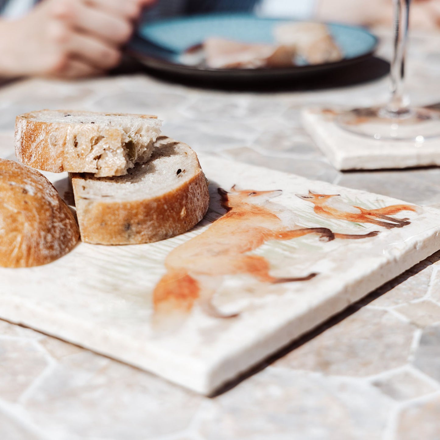 A garden table set for al fresco dining with marble platters, including a medium square marble platter used as a breadboard, featuring a fox and fox cub watercolour design.