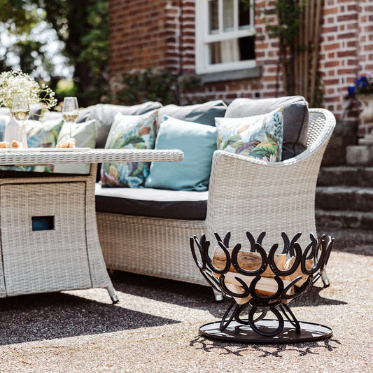 A handmade horseshoe fire pit next to a seating area in the garden of a country house. The fire pit has been filled with firewood and has a black painted finish.