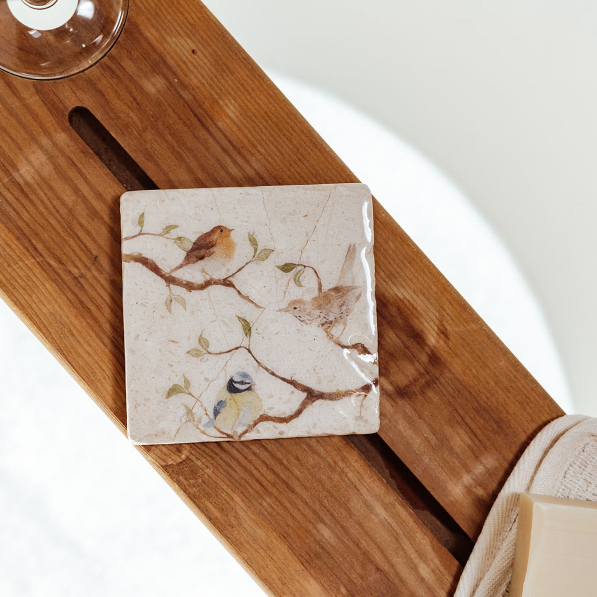 A square marble coaster on a wooden bath tidy above a bubble bath. The coaster features a British garden birds watercolour design.