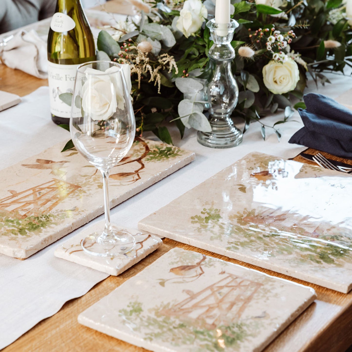 A wooden dining table set with cream marble placemats and coasters. The placemats are square and feature bees and a beehive in a buttercup meadow in a watercolour style.