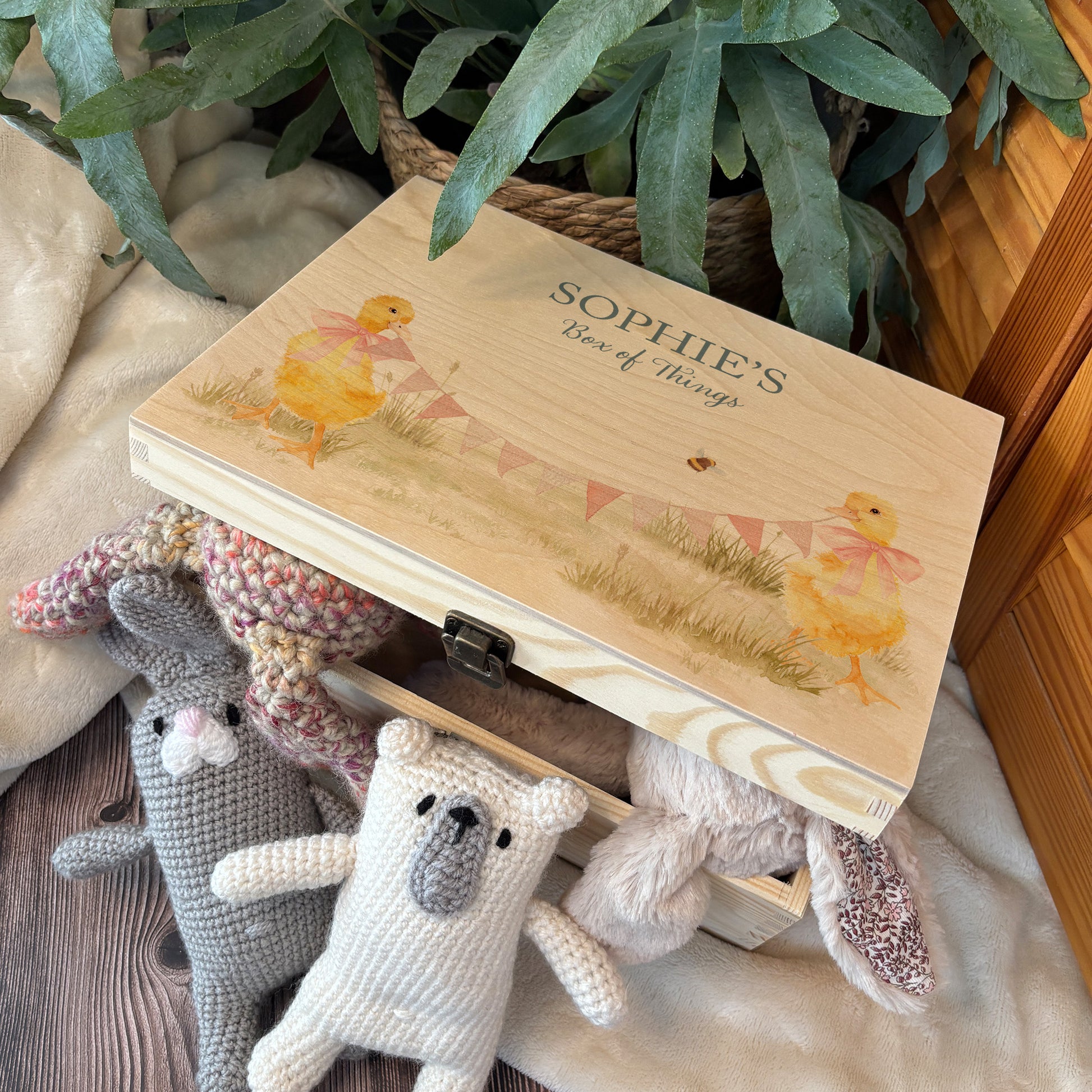 Medium sized wooden pine personalised keepsake box. The design theme is perfect for New babies, Welcome to the world, baby showers and christenings. This design has baby ducklings  in meadow style grass and a buzzy Bee flying around. The picture also shows an example in a nursey room filled with nappies, vests and teddys.