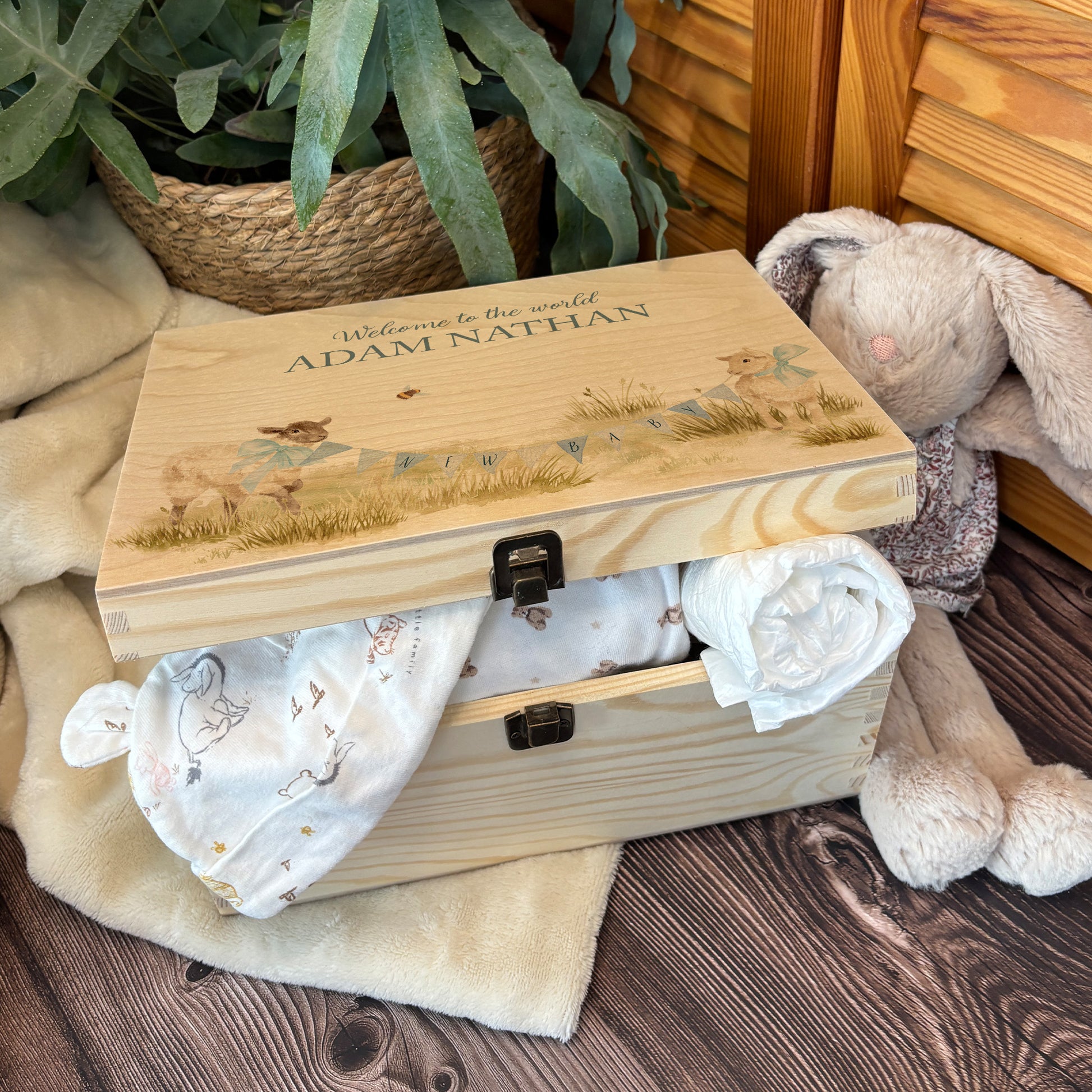 Medium sized wooden pine personalised keepsake box. The design theme is perfect for New babies, Welcome to the world, baby showers and christenings. This design has baby lambs  in playing in grass with a little bee flying around.  This design can be personalised by adding a beautiful bunting and a luxurious bow around their necks in blue or pink. you can also add names , The picture also shows an example in a nursey room filled with nappies, vests and teddys.