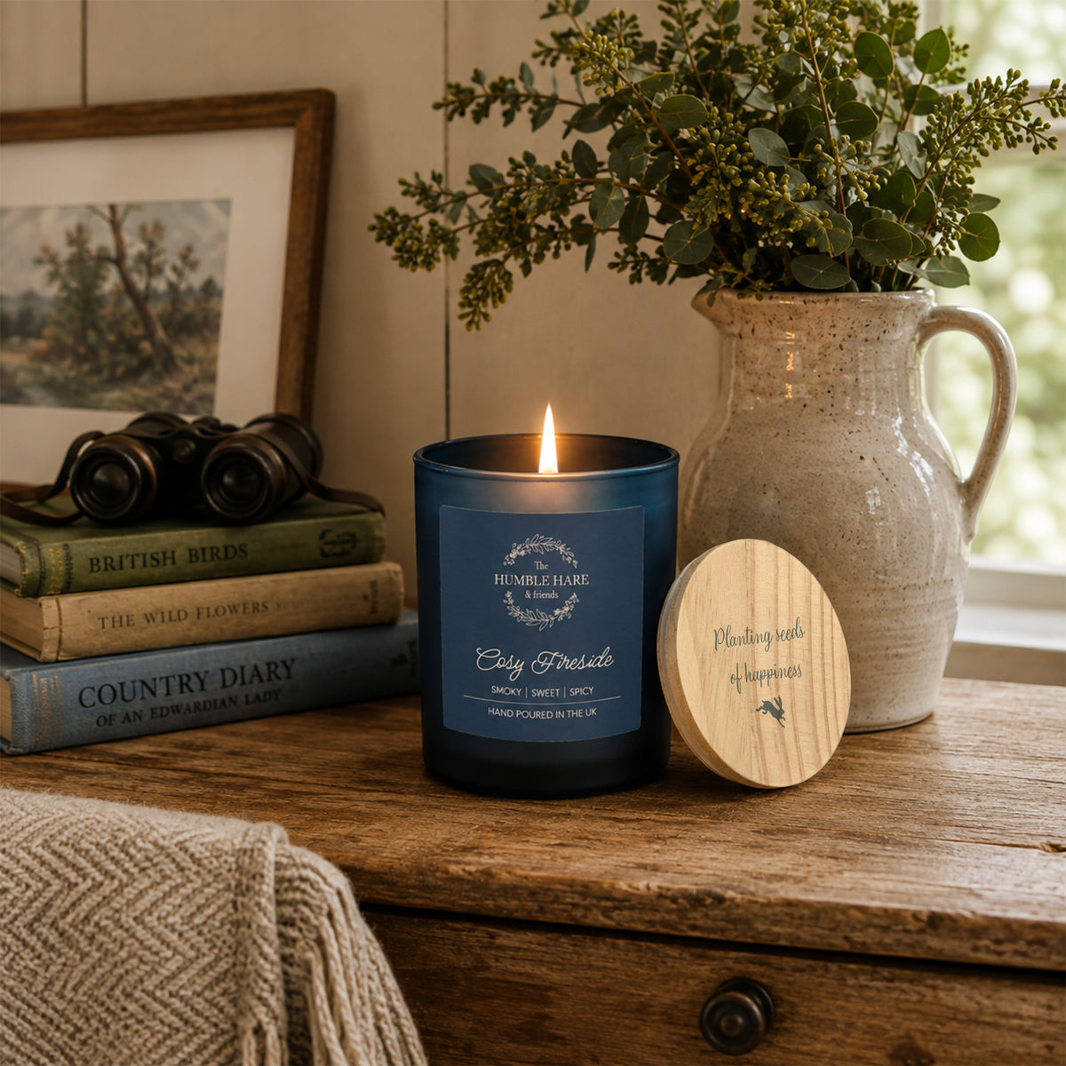 A dark blue glass jar candle with a light natural wood lid with the quote 'Planting seeds of happiness' and a small leaping hare silhouette.