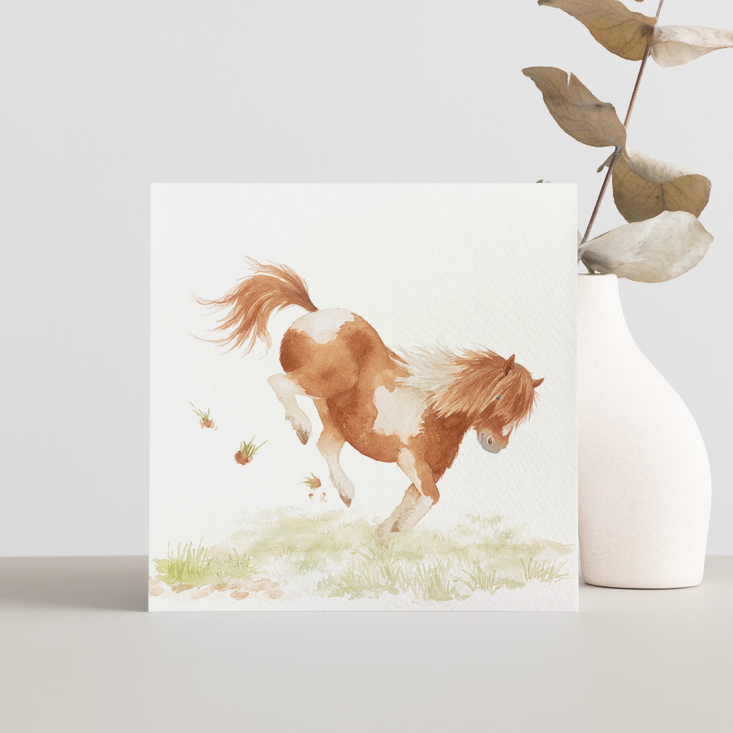 A square greetings card with a watercolour design of a bucking Shetland pony in a field.