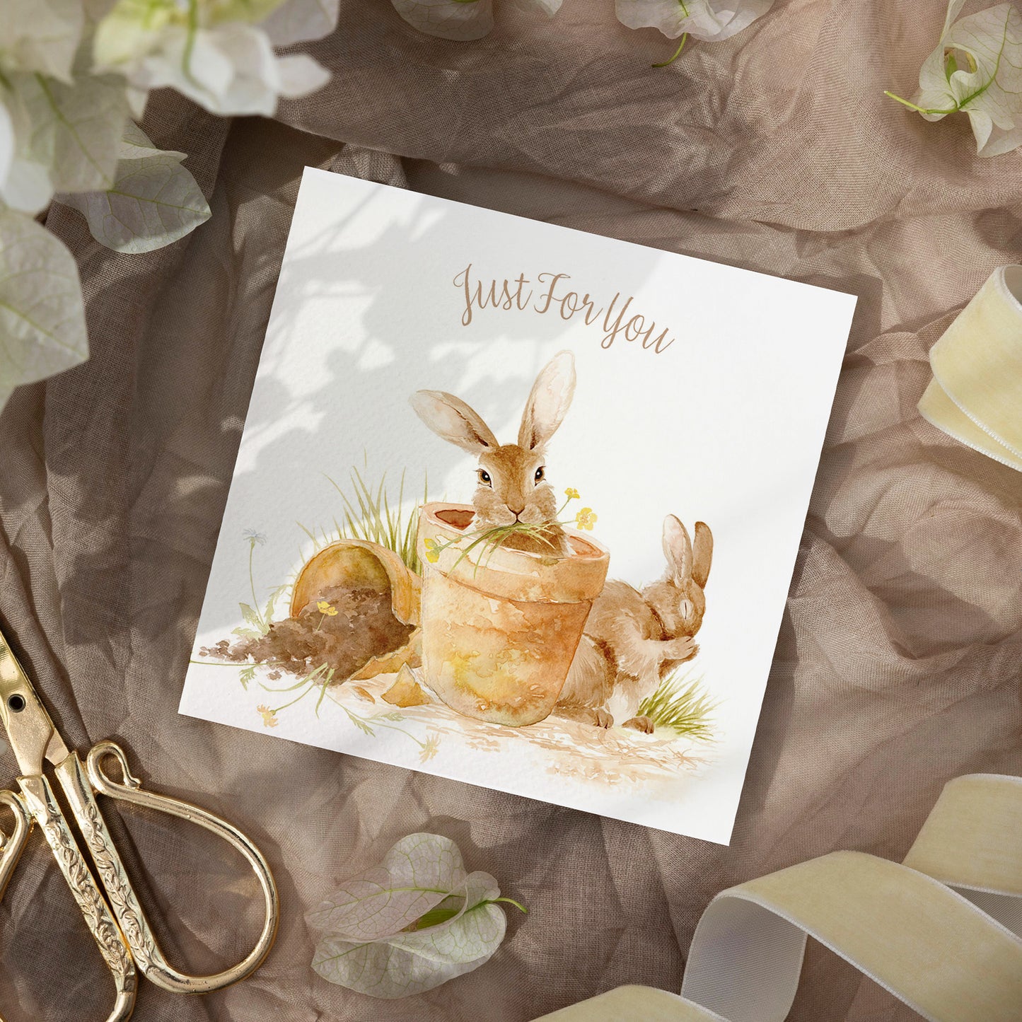 A square greetings card with a watercolour design of rabbits in flowerpots eating flowers. The Card reads 'Just For You'. The card is laid on neutral colour fabric with ribbons and scissors.