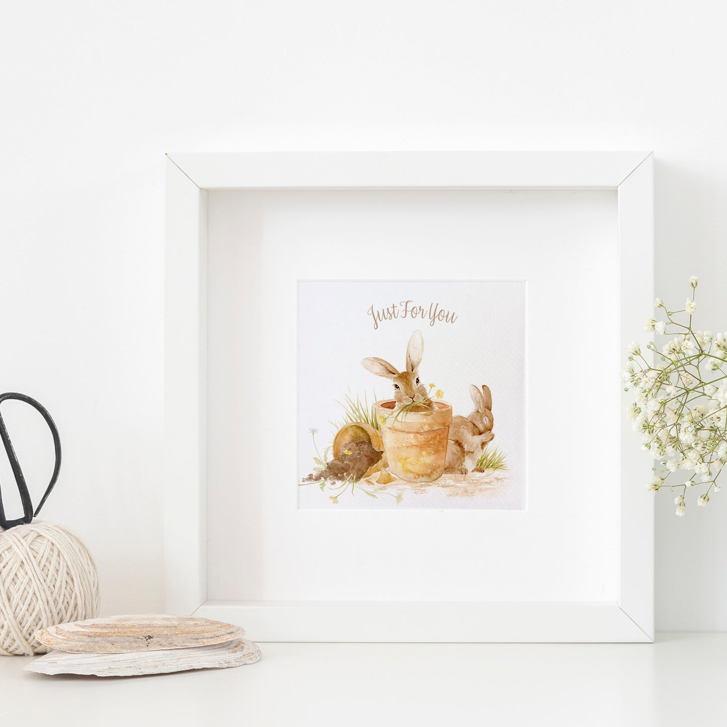 A framed greetings card with a watercolour design of rabbits in flowerpots eating flowers. The Card reads 'Just For You'.