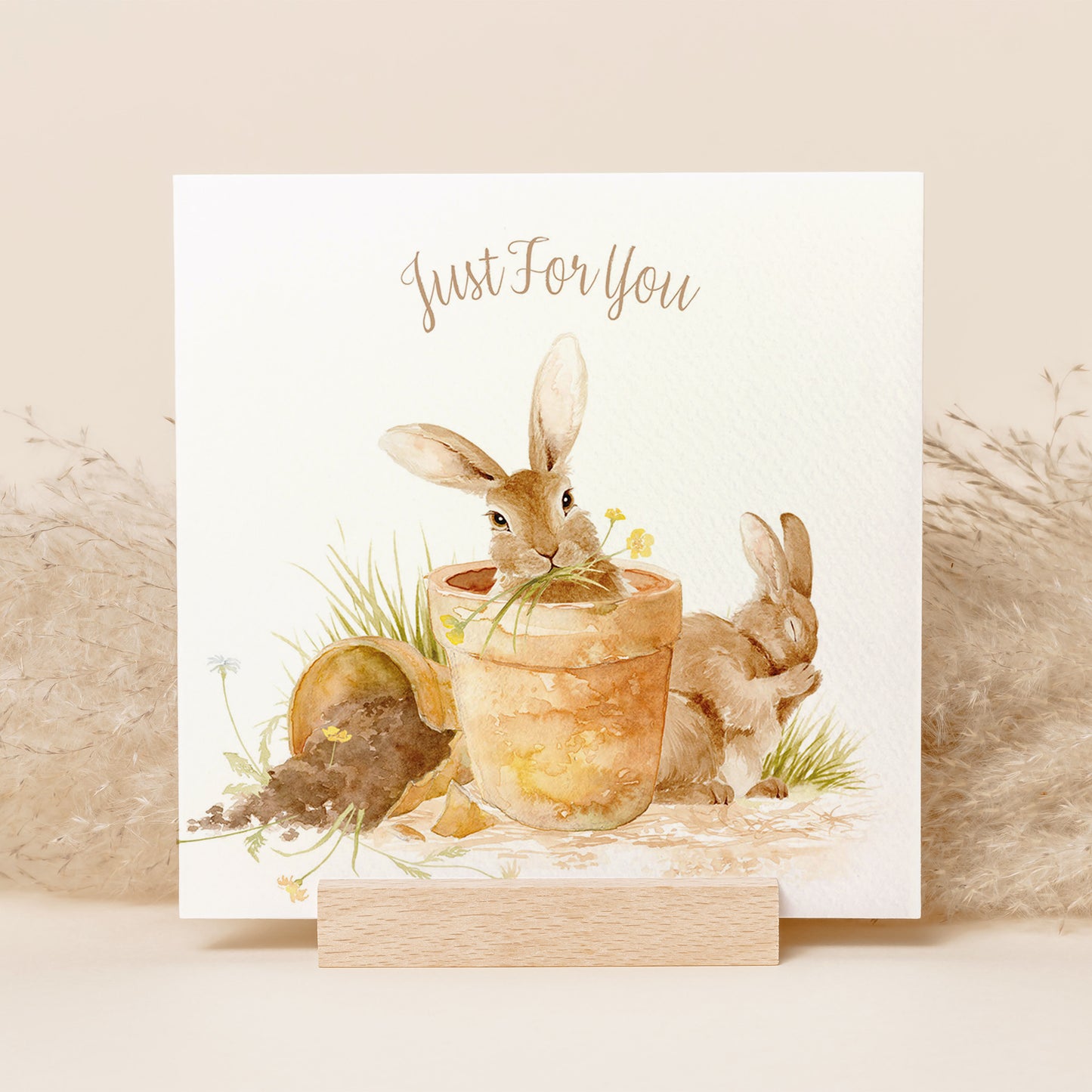 A square greetings card with a watercolour design of rabbits in flowerpots eating flowers. The Card reads 'Just For You'.