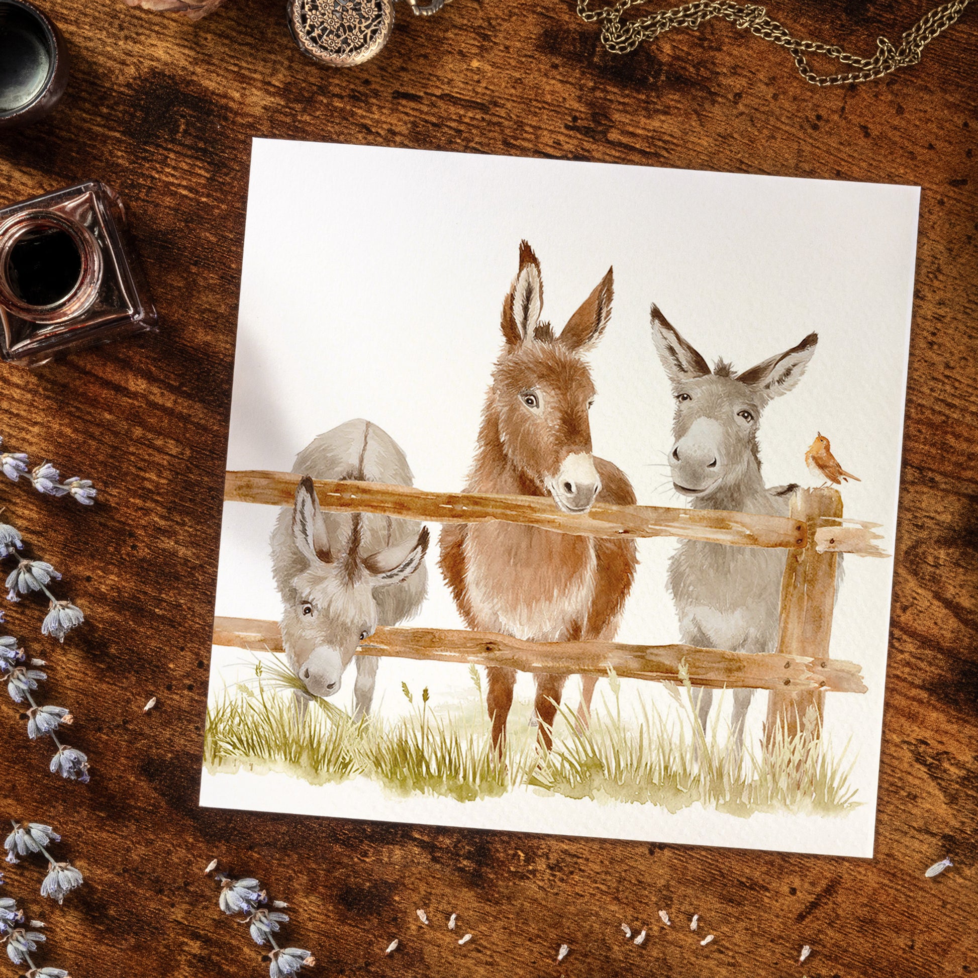 A bookmark greetings card featuring a watercolour design of three donkeys peering over a wooden fence. The card is laid on a wooden desk surrounded by stationery.