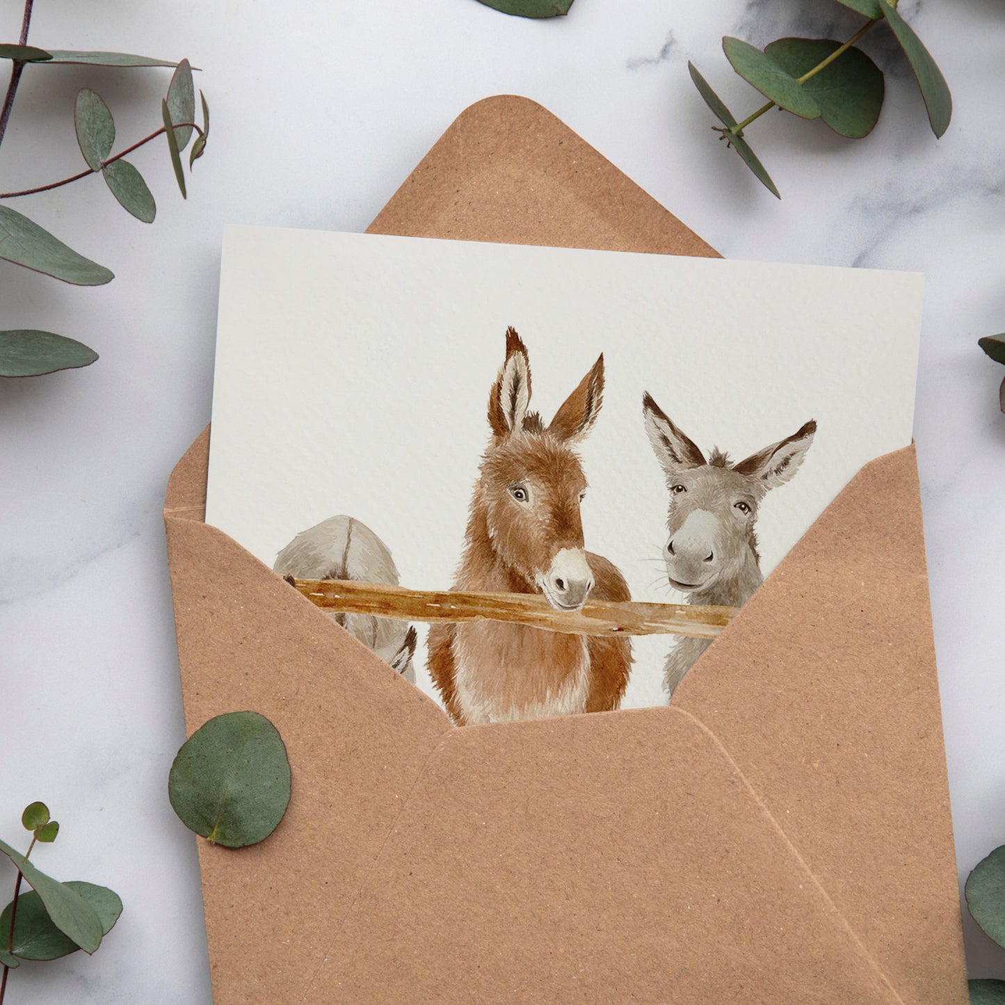 A bookmark greetings card with a watercolour scene of three donkeys peering over a wooden fence. The card is inside a brown kraft envelope.