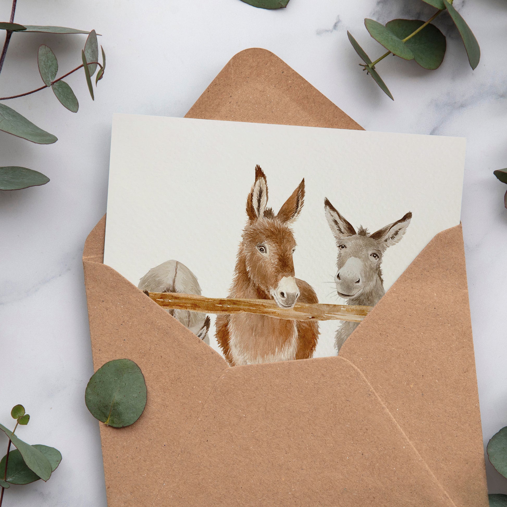 A bookmark greetings card with a watercolour scene of three donkeys peering over a wooden fence. The card is inside a brown kraft envelope.