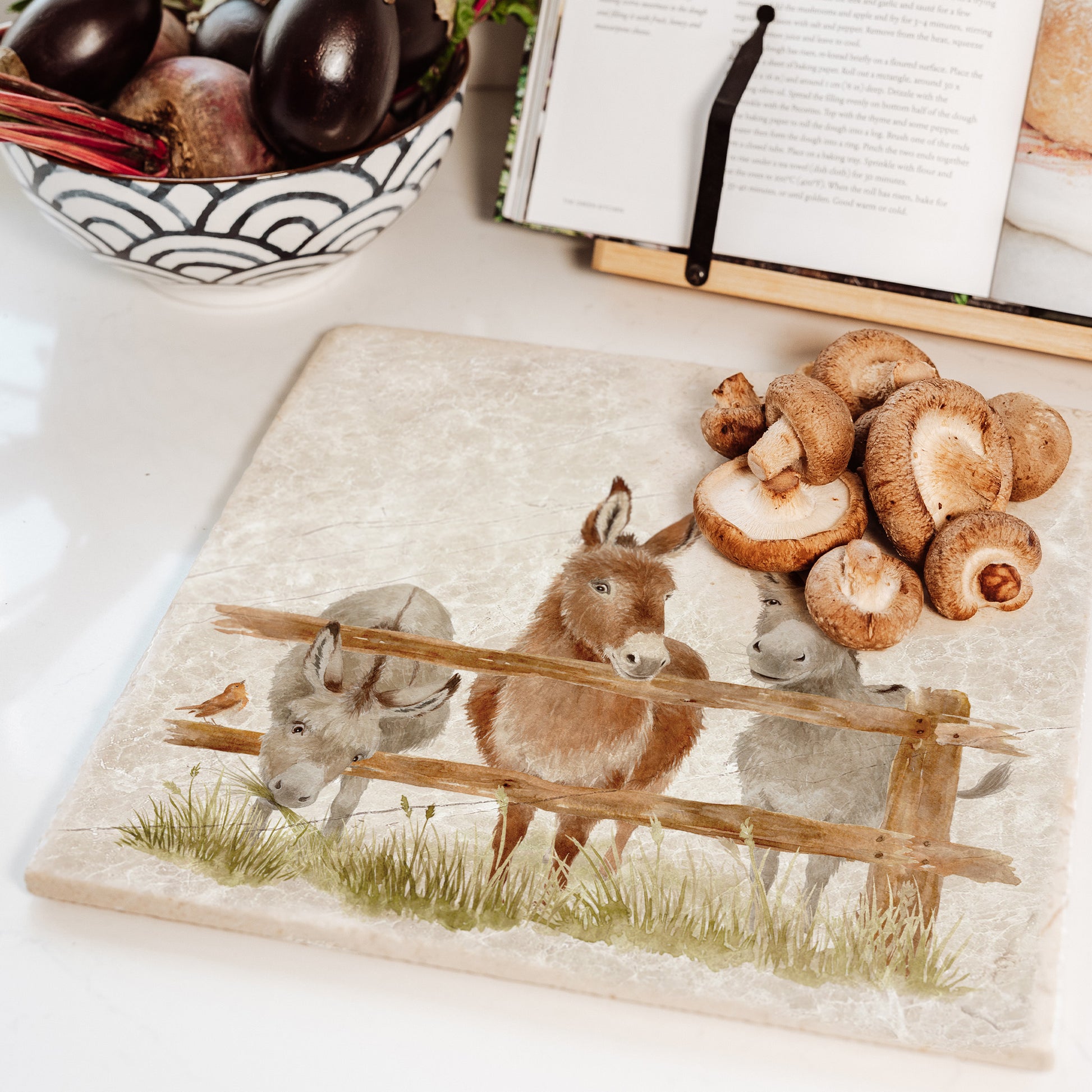 A large square marble worktop saver featuring three donkeys peeking over a wooden fence. The platter is set on a kitchen worktop.