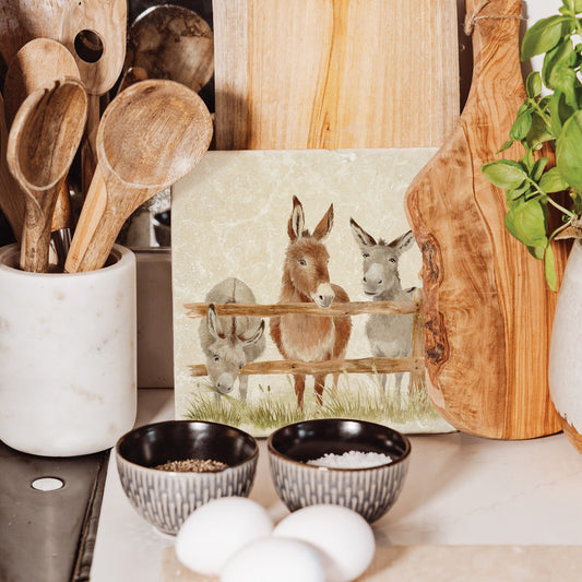A handmade medium marble platter featuring a watercolour design of three donkeys at a fence. The marble platter is leant up against a kitchen worktop with other wooden serving boards.