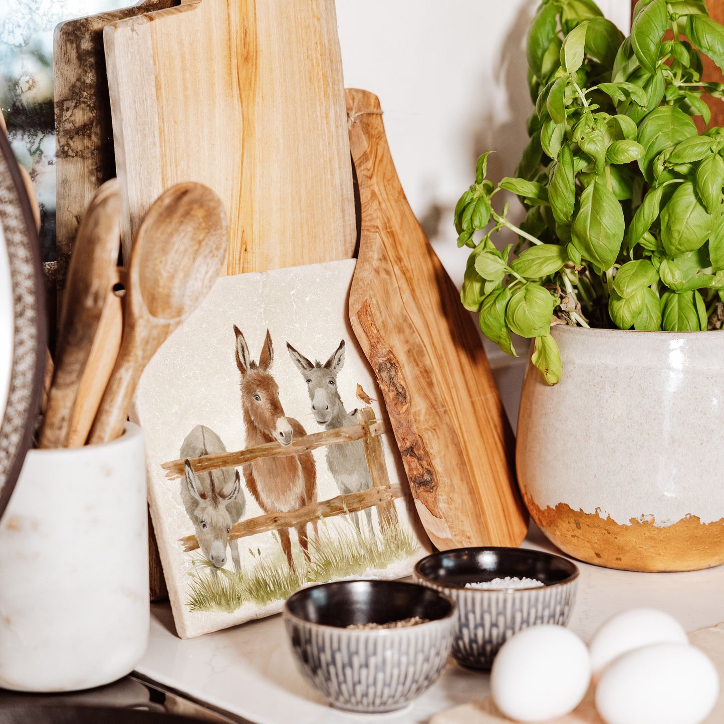 A medium marble platter featuring a watercolour design of three donkeys at a fence. The marble platter is leant up against a kitchen worktop with other wooden cheeseboards.