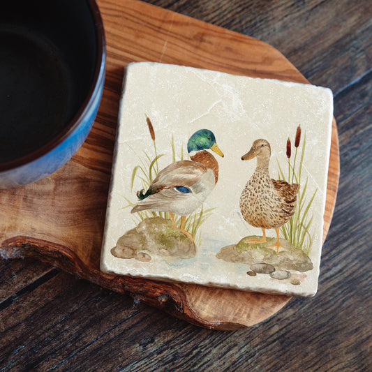 A square tumbled marble coaster featuring a watercolour design of two mallard ducks standing at the edge of a pond. The coaster is set on a kitchen worksurface.