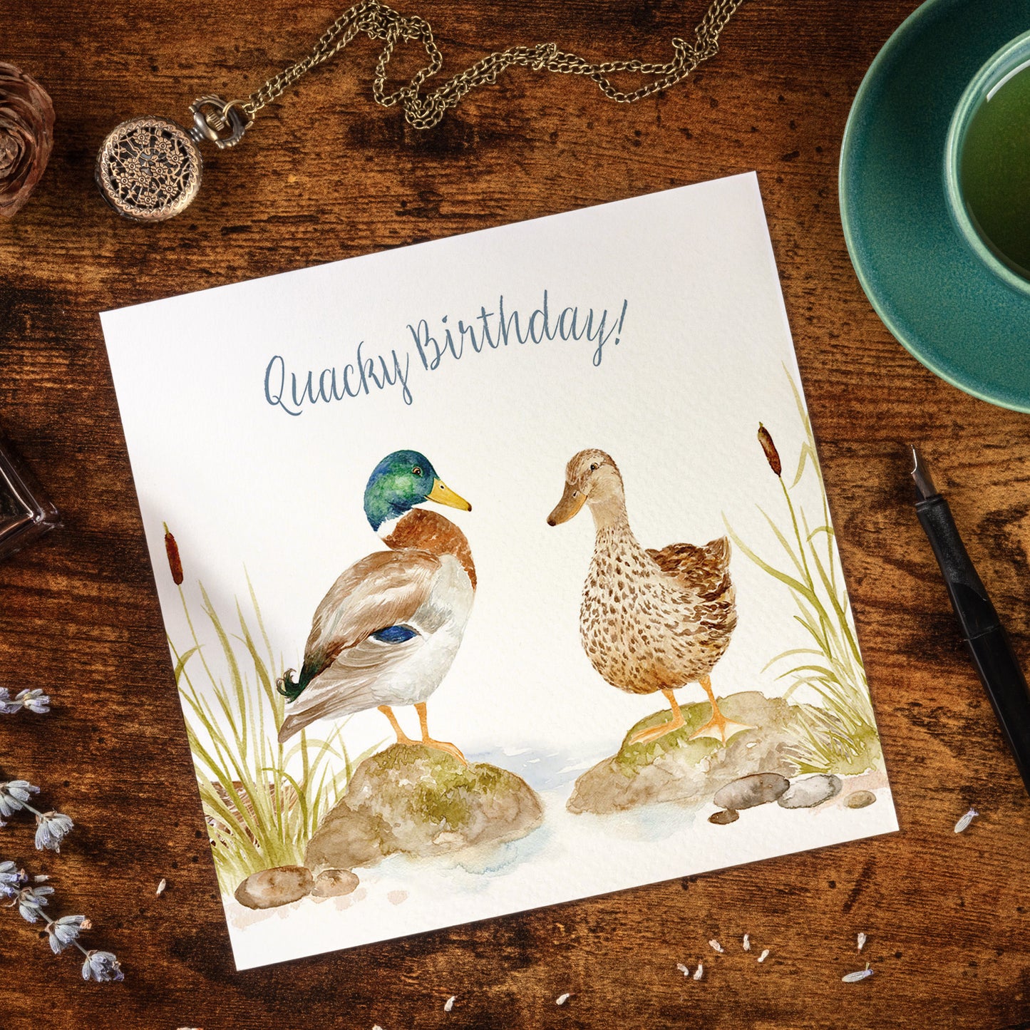 A square greetings card featuring a watercolour design of two Mallard ducks that reads 'Quacky Birthday'. The card is laid on a wooden desk surrounded by stationery items.