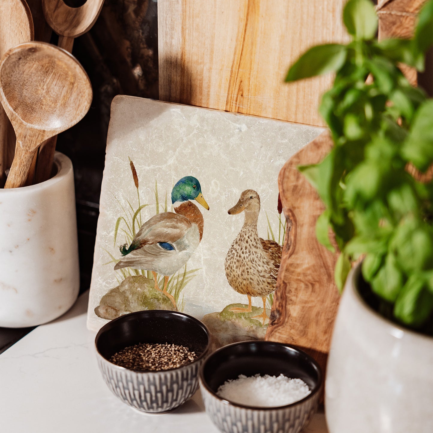 A square medium marble platter featuring a watercolour scene of mallard ducks at a pond. The platter is propped up on a kitchen worksurface next to the hob.