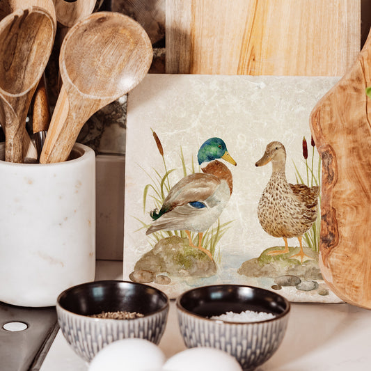 A medium marble cheeseboard featuring a watercolour scene of mallard ducks at a pond. The platter is propped up on a kitchen worksurface.