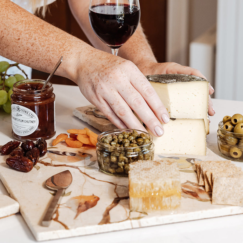 A large marble serving platter with a watercolour design featuring British garden birds on branches. The platter is being used as a cheeseboard, with cheese, crackers, honeycomb and chutney arranged on it.