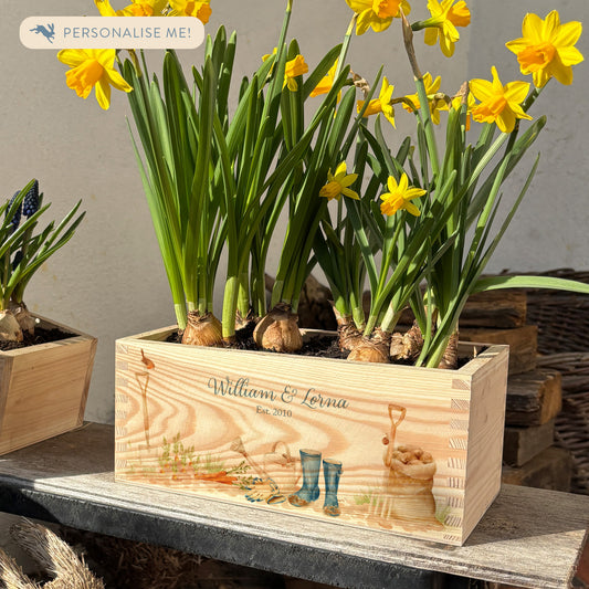 A rectangle wooden planter full of bright daffodils. The planter box shows a watercolour design of a gardening scene with welly boots, watering can, sack of potatoes, carrots, robin, and garden spade and fork. The planter is personalised with a couple's names and Est. on date. The perfect gift for weddings, anniversaries, and engagements, or other special occasions.