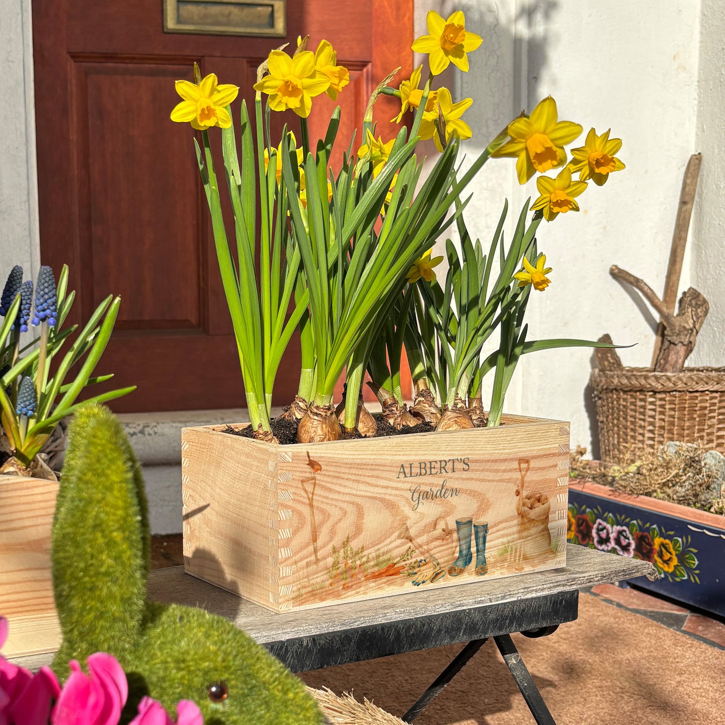 A rectangle wooden planter full of bright daffodils. The planter box shows a watercolour gardening scene with welly boots, watering can, garden spade and fork, and a sack of potatoes. The planter is personalised with a special message and custom name. An ideal gift for gardeners, birthdays, new home, wedding, anniversary, or other special occasions.