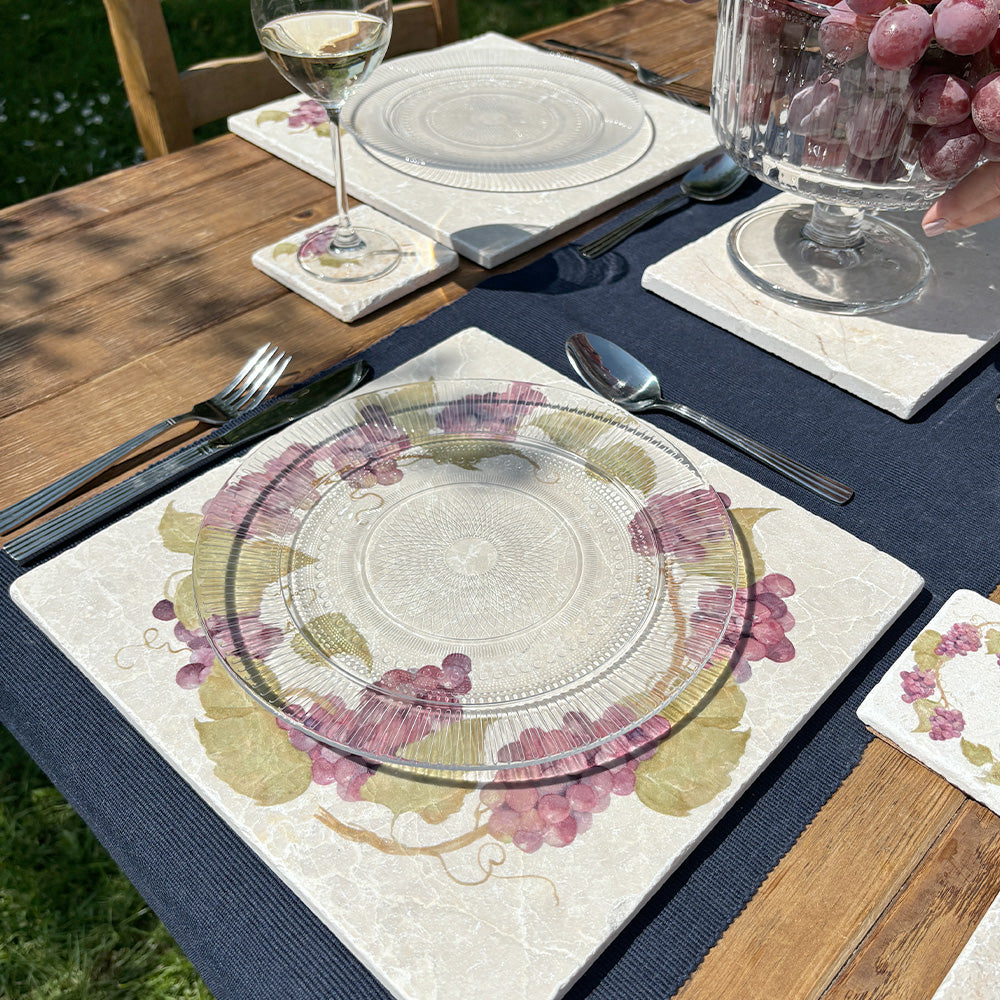 A garden table in the sunshine, the table is set with marble placemats and coasters featuring a rustic grape vine wreath design.
