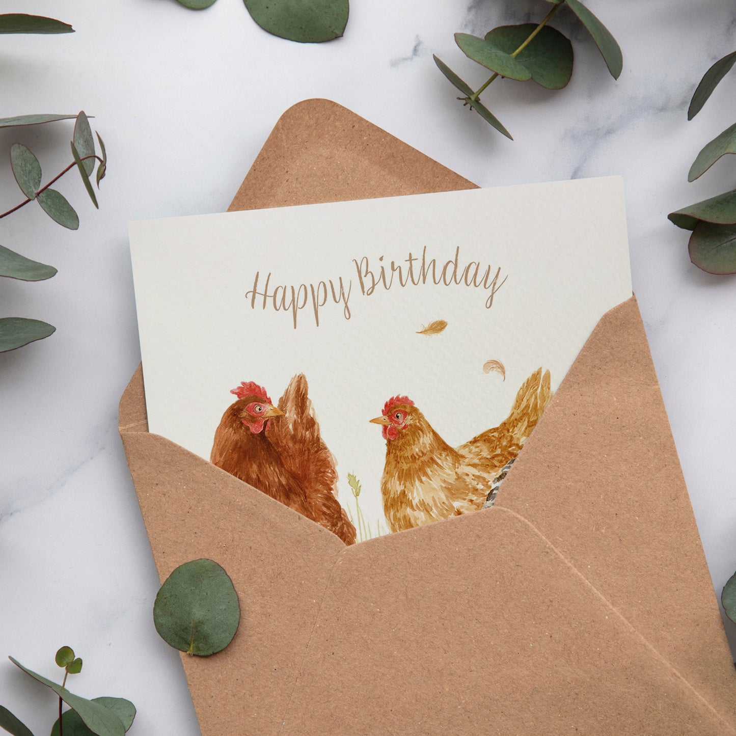 A bookmark greetings card with a watercolour scene of three hens that reads 'Happy Birthday'. The card is inside a brown kraft envelope.