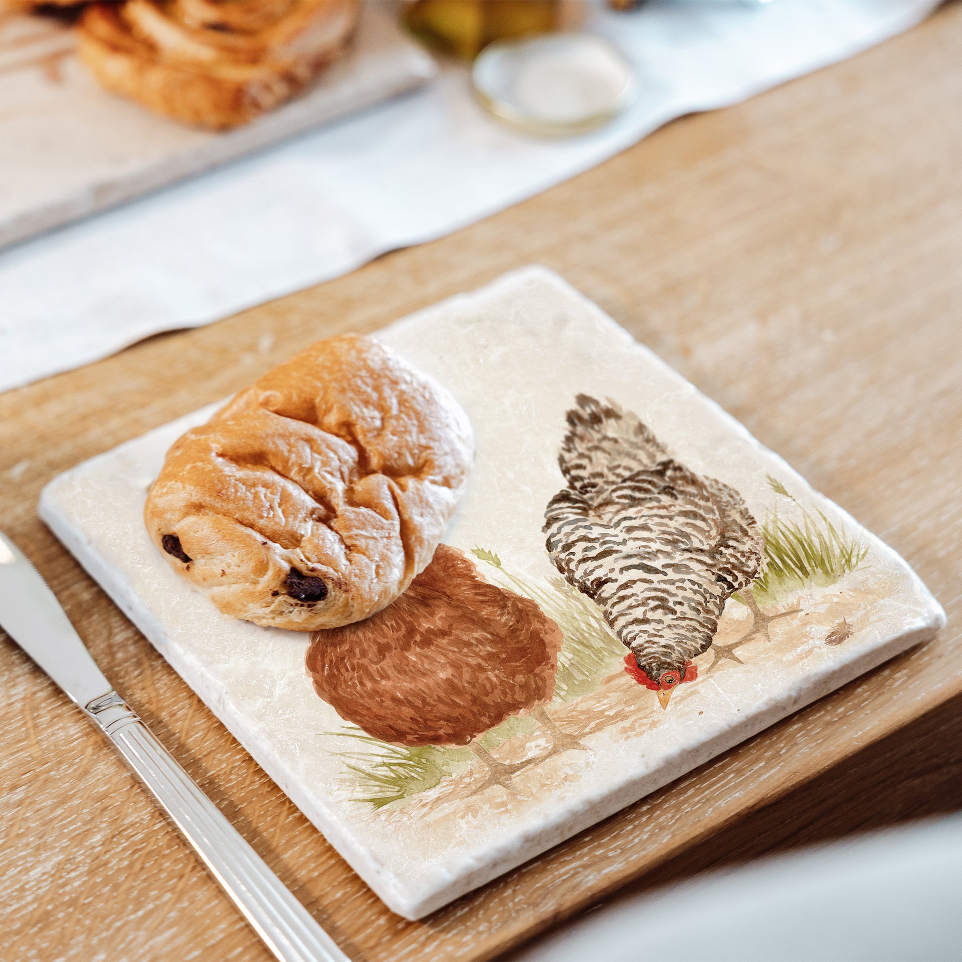 A medium marble platter set on a farmhouse kitchen table and used to serve pastries. The platter features a watercolour design of two hens.