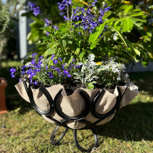 A handmade ironmongery planter made from recycled horseshoes. The garden planter is set on a lawn and filled with flowers and shrubs.