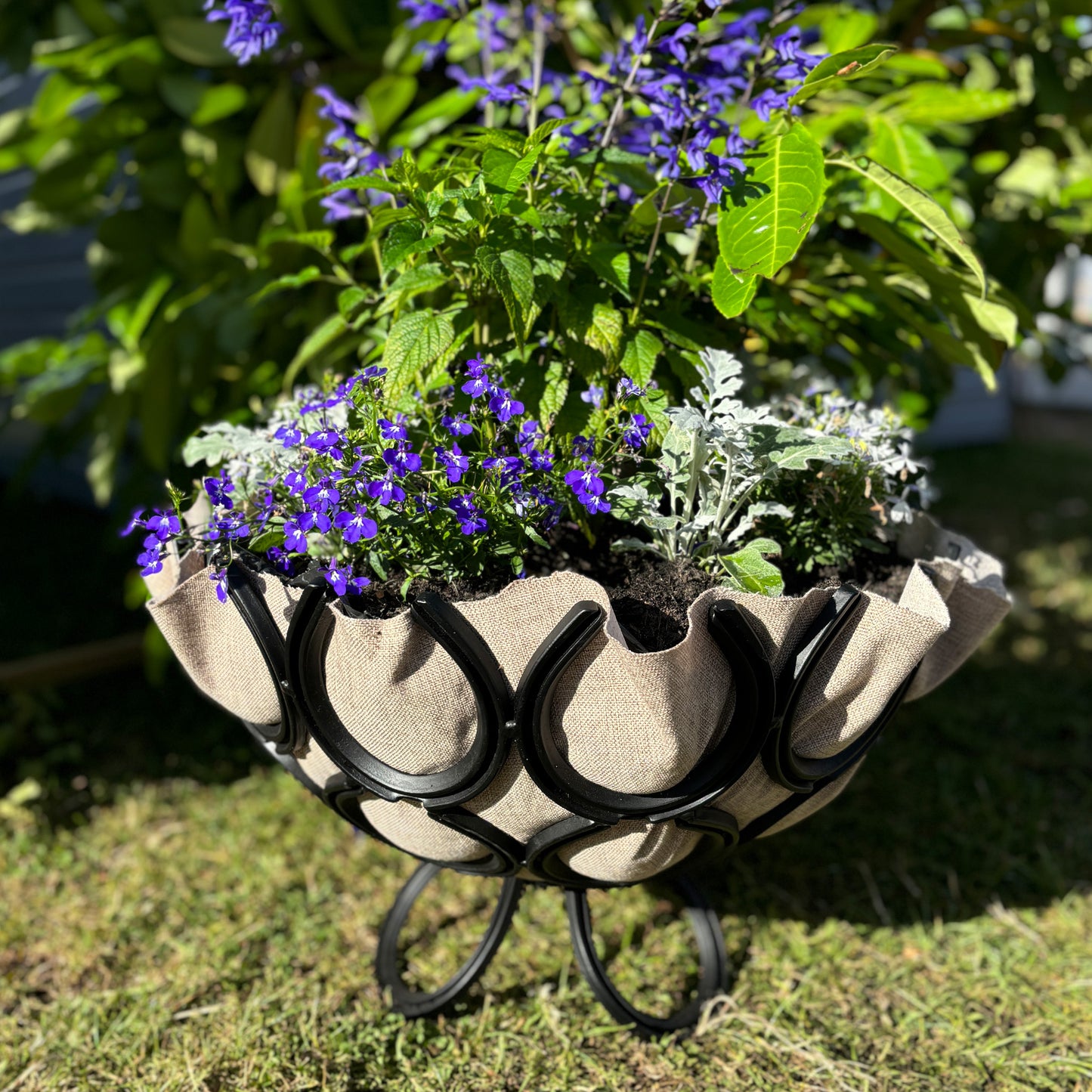 A handmade ironmongery planter made from recycled horseshoes. The garden planter is set on a lawn and filled with flowers and shrubs.