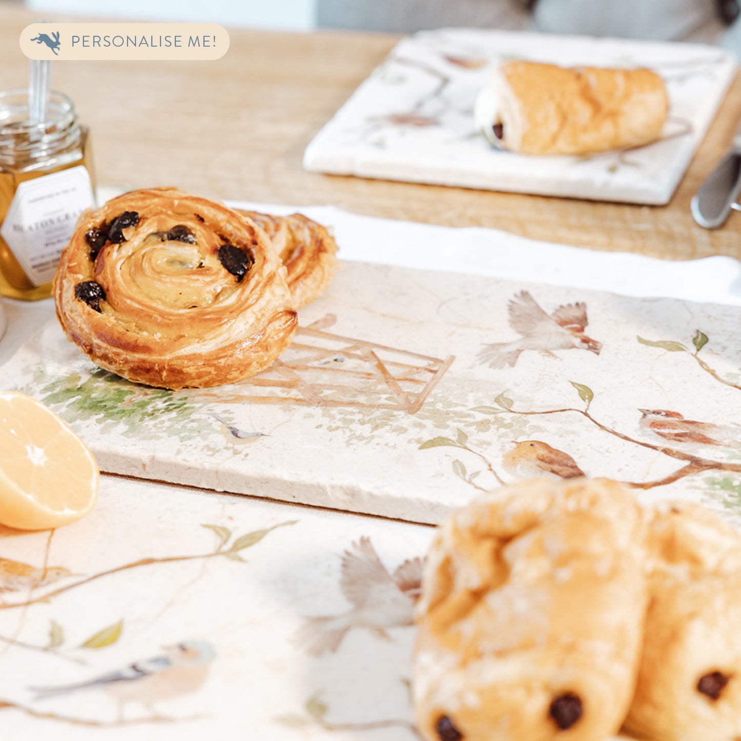 A breakfast table set with a marble sharing platter featuring British garden birds in the hedgerow around the paddock gate, in a watercolour style. The platter is being used to serve pastries and fruit.