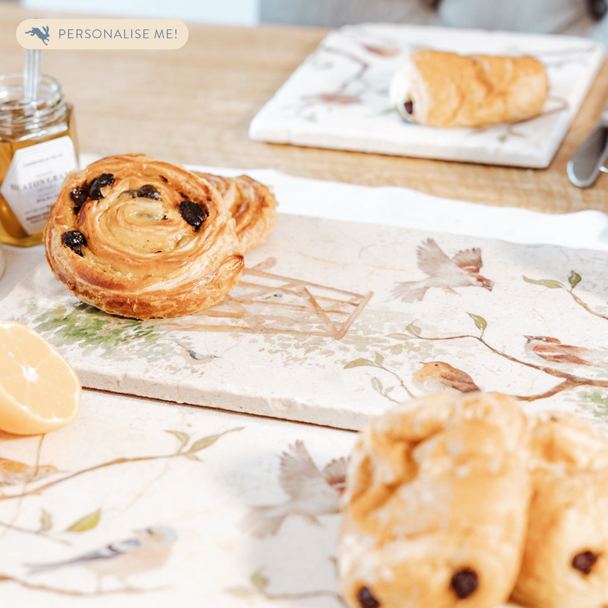 A breakfast table set with a marble sharing platter featuring British garden birds in the hedgerow around the paddock gate, in a watercolour style. The platter is being used to serve pastries and fruit.