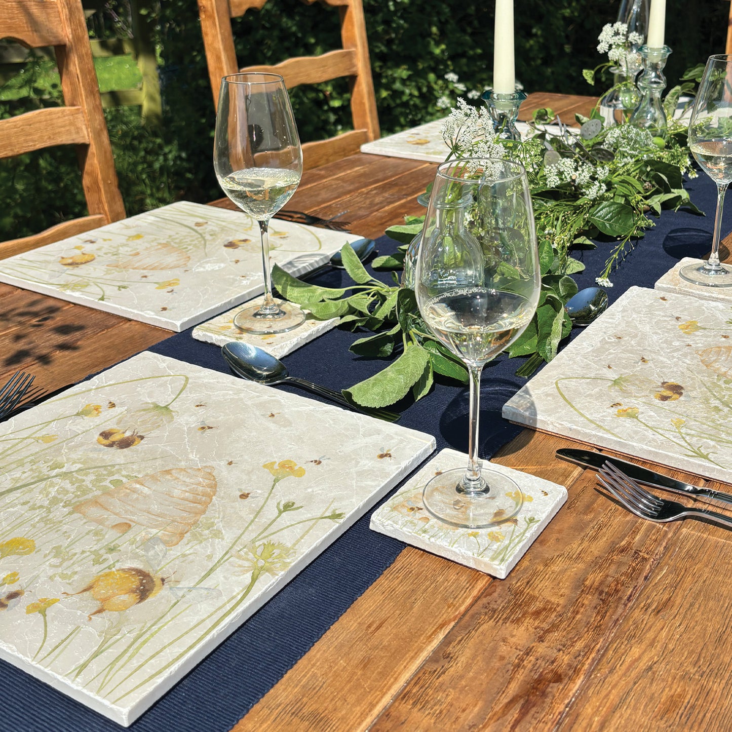 A wooden farmhouse dining table set with large square marble placemats. The placemats feature bees and a beehive in a buttercup meadow, in a watercolour style.