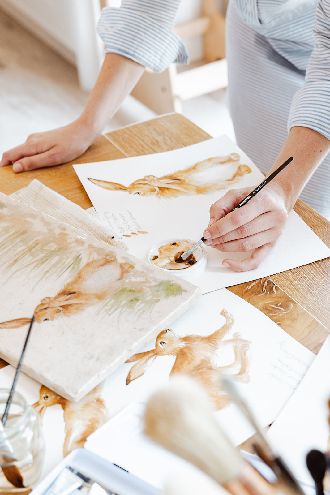 An artist painting hares in different poses, using watercolour paints on paper.