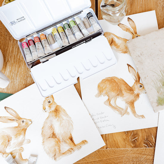 Art supplies including watercolour paints and paintings of a hares on a wooden table.