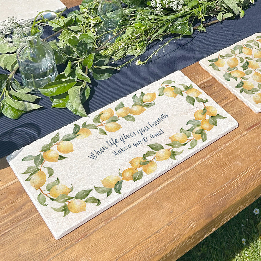 A personalised marble sharing board featuring a wreath of lemons and leaves, with a bespoke message in the centre of the design reading 'When life gives you lemons make a gin & tonic!'. The custom sharing platter is placed on a sunny garden table ready for an al fresco lunch or dinner.