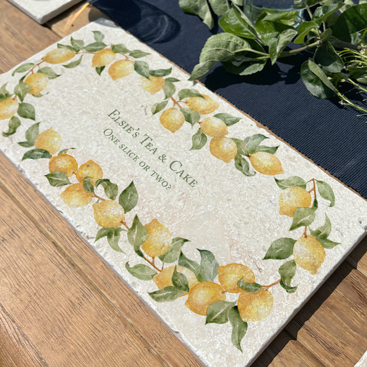 A personalised marble sharing board featuring a wreath of lemons and leaves, with a bespoke message in thye centre of the design reading 'Elsie's Tea & Cake, One Slice or Two?'. The custom sharing platter is placed on a sunny garden table ready for an al fresco lunch or dinner.