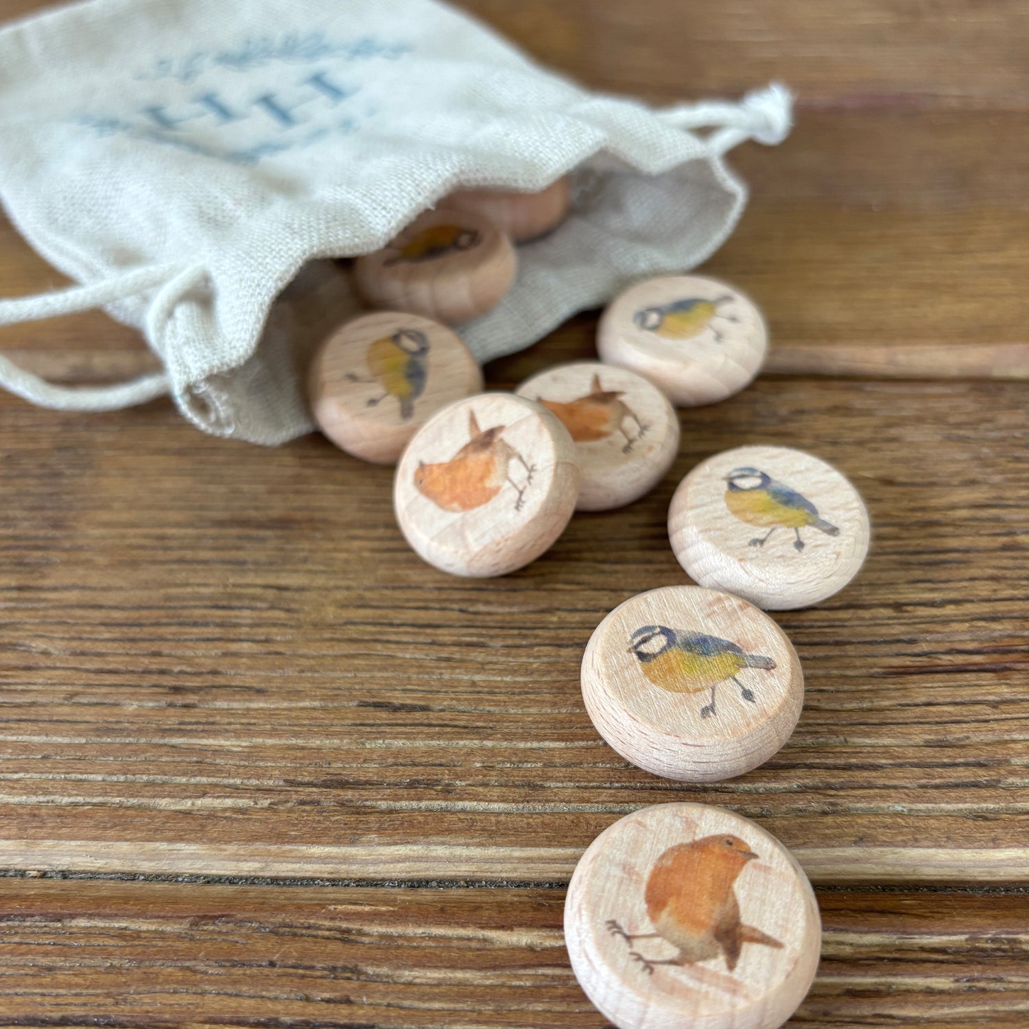 A small cotton bag full of wooden draughts or checkers counters, each featuring a watercolour illustration of a British bird, such as robins and blue tits.