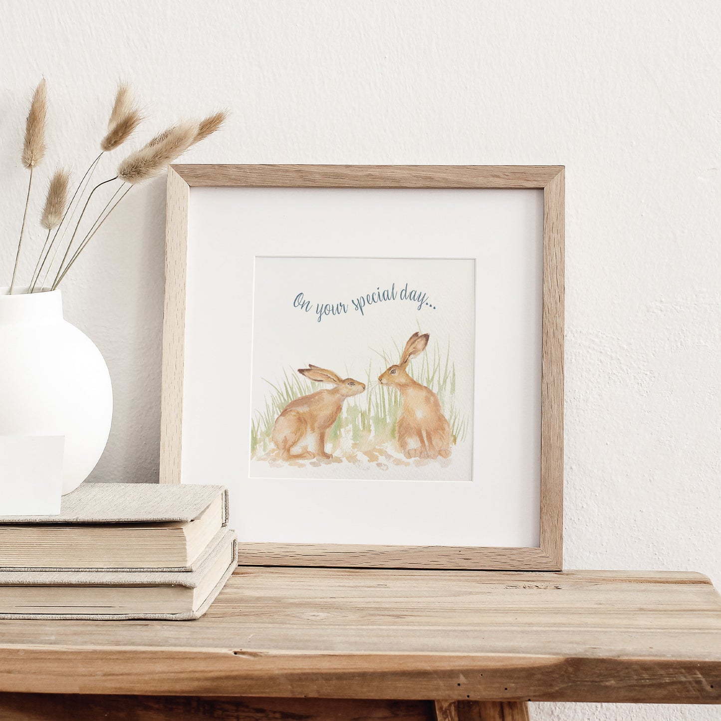 A greetings card displayed as an art print in a neutral coloured frame propped up on a bookshelf. The card reads On Your Special Day in dark blue text above a hare couple in a watercolour style.