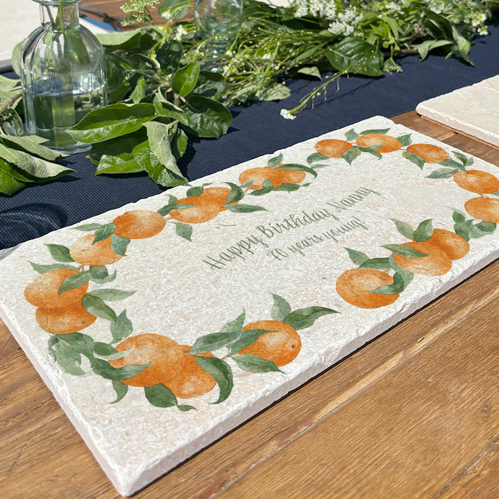 A personalised marble sharing board featuring a wreath of oranges and leaves, with a bespoke message in the centre of the design. The personalised marble platter is placed on a sunny outdoor table.