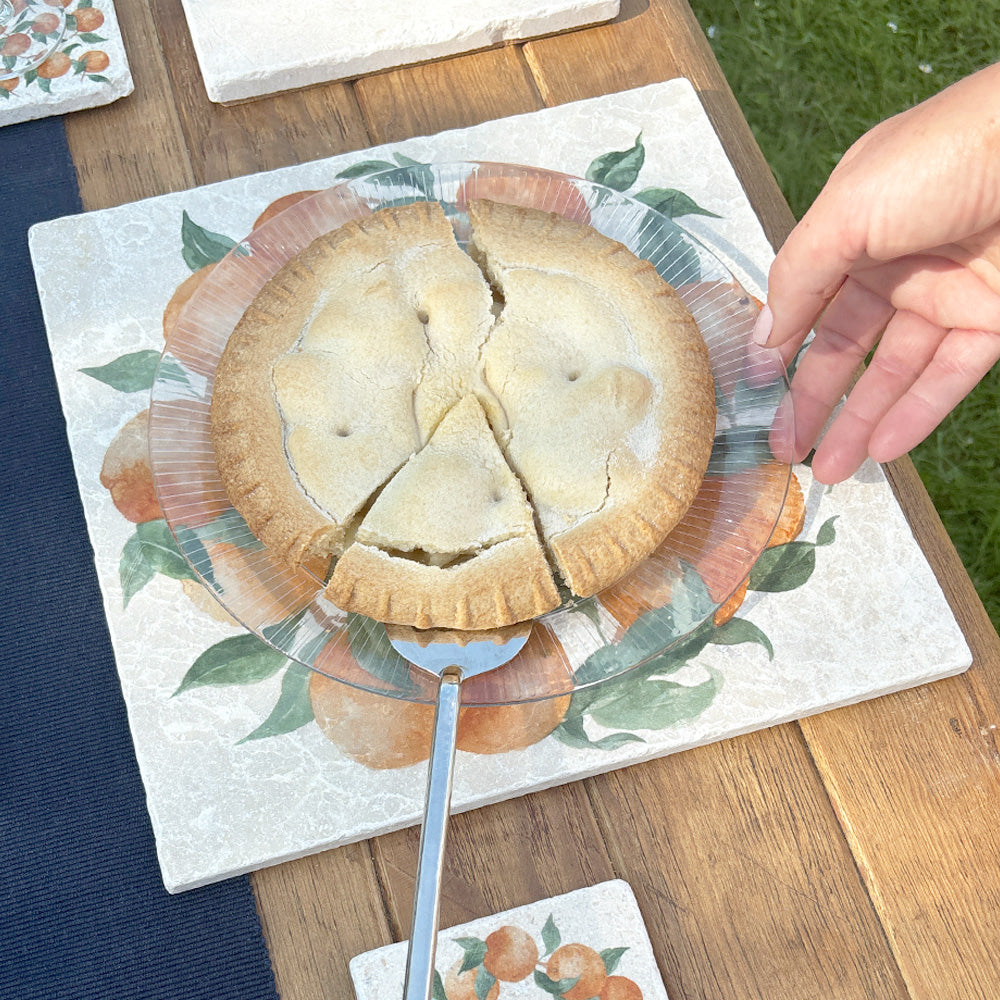 A large marble platter featuring a watercolour orange wreath design. The platter is being used as a trivet to serve a delicious apple pie.