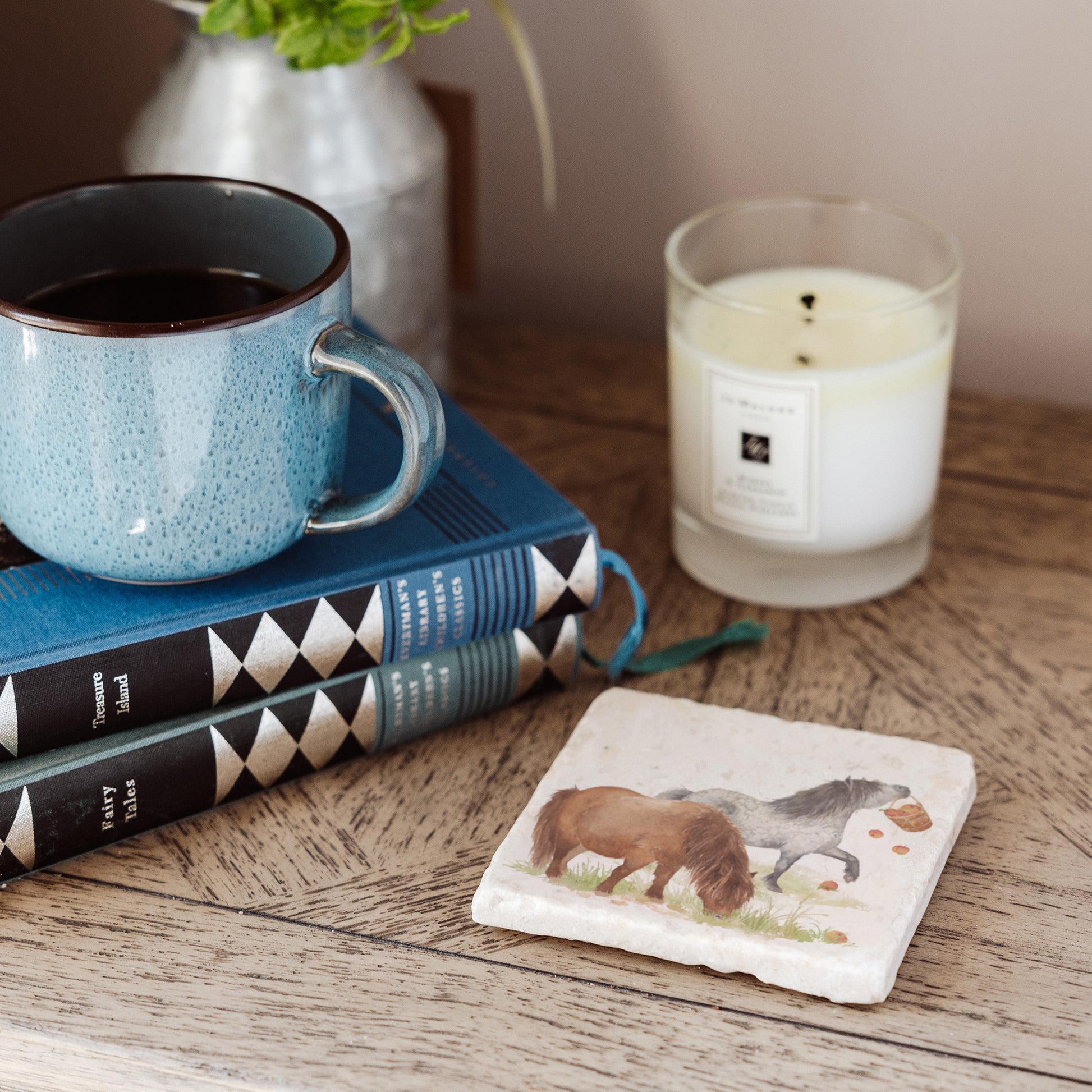 A square marble coaster placed on a bedside table, featuring a watercolour design of two cheeky Shetland ponies stealing apples.