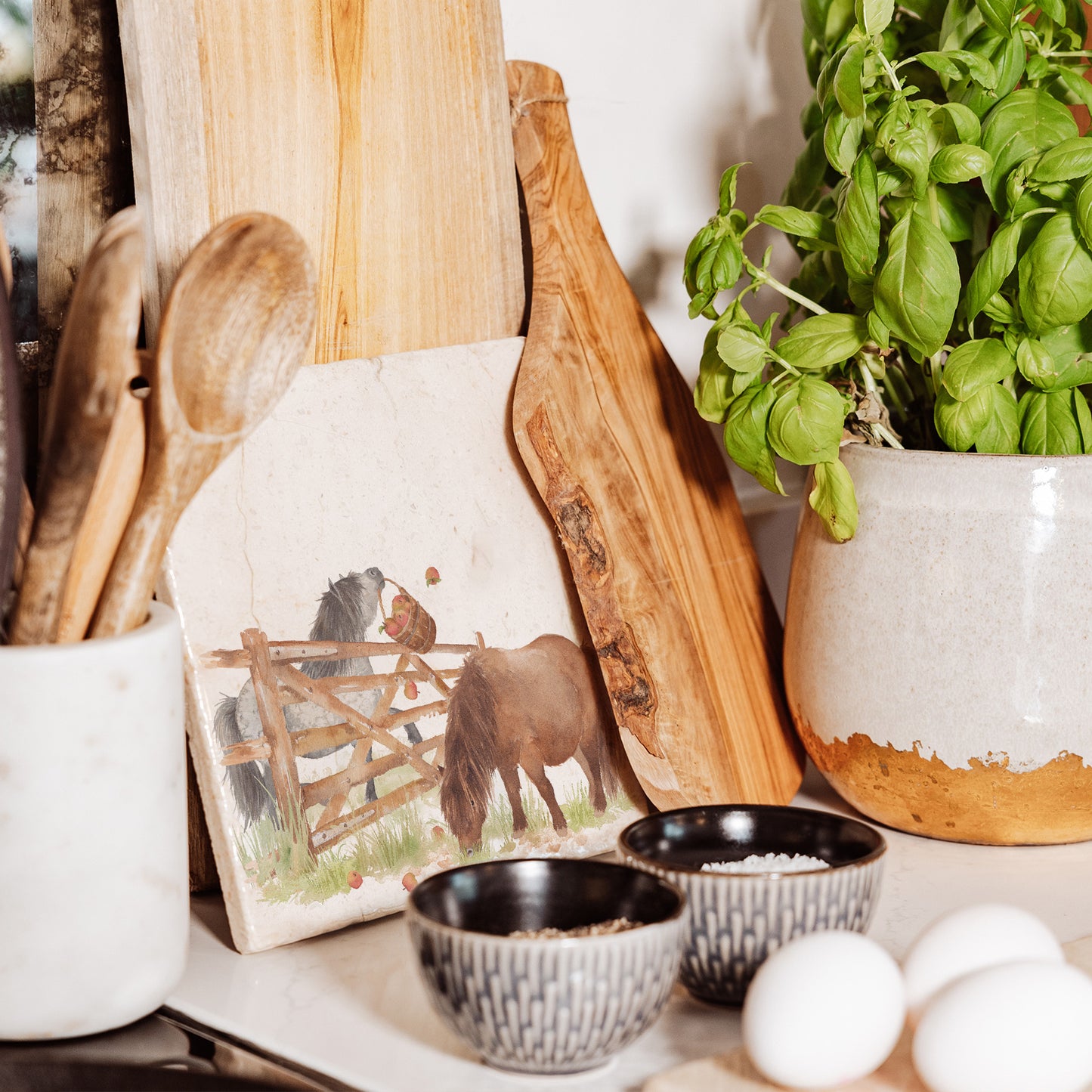 A medium square multipurpose marble chopping board propped up on a country kitchen worksurface, featuring a watercolour design of two cheeky Shetland ponies stealing apples.