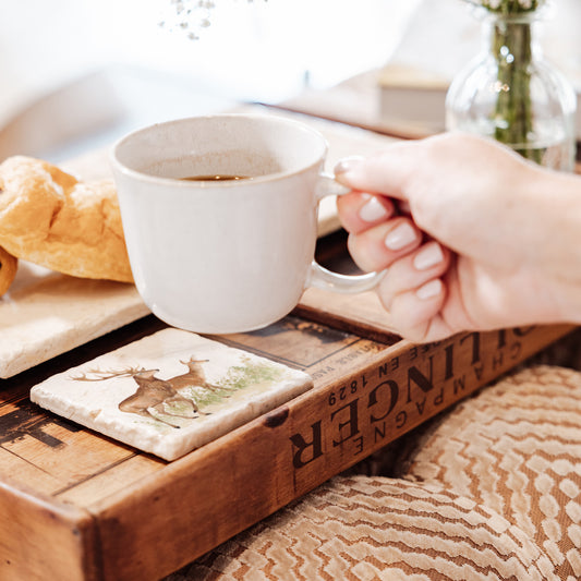 A woman's hand placing a coffee cup on a marble coaster. The coaster design features a red deer stag and hind.