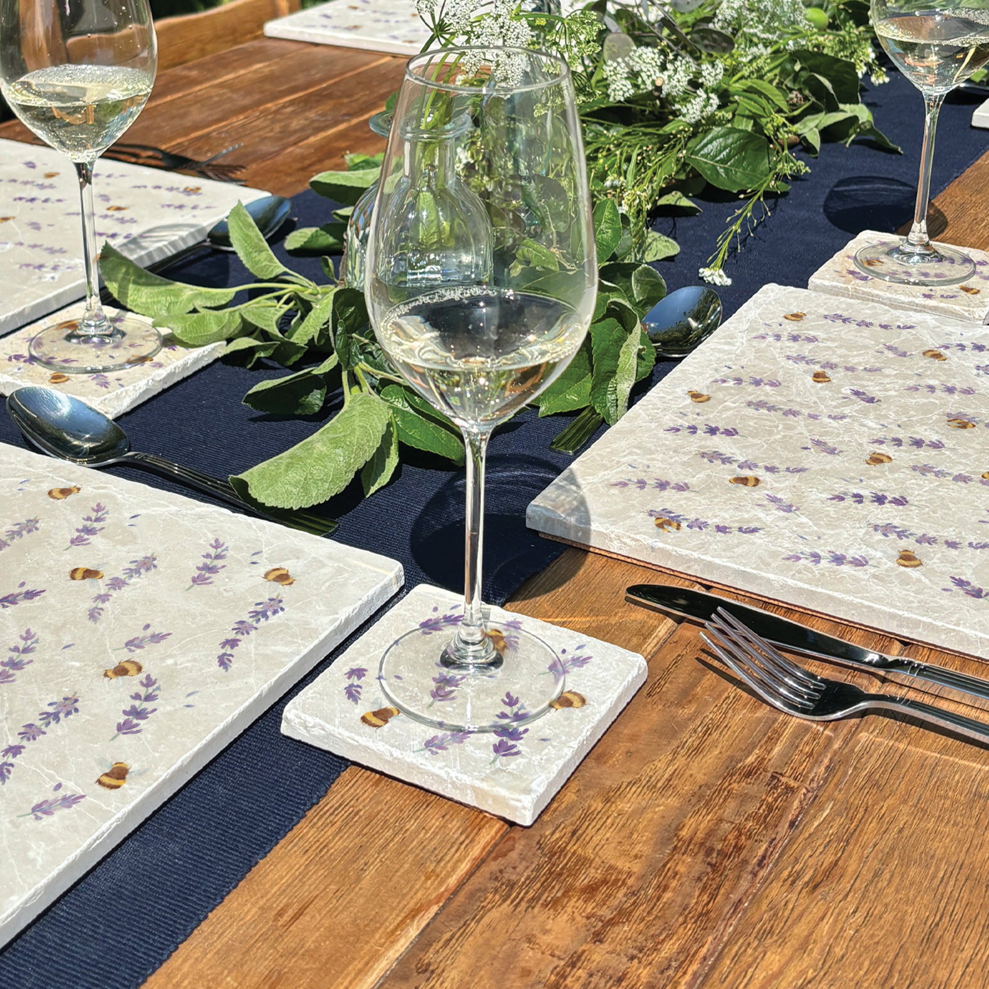 A square marble coaster with a watercolour pattern of busy bees and purple lavender flowers. The coaster is set on an outdoor table with matching placemats.