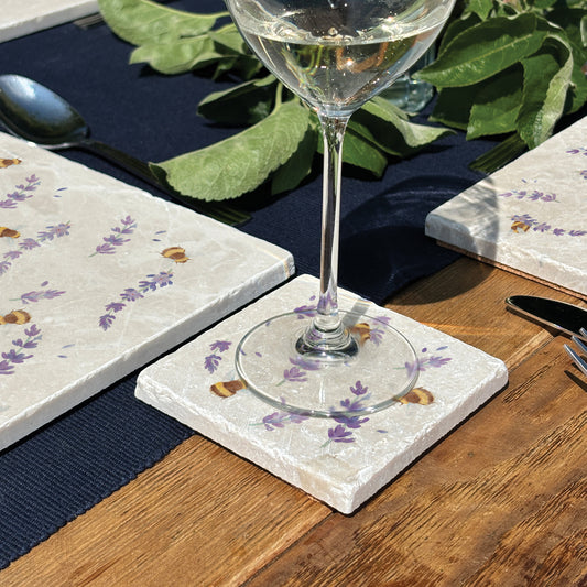 A handmade marble coaster featuring a watercolour pattern of busy bees and lavender flowers. The coaster is set on a garden dining table.