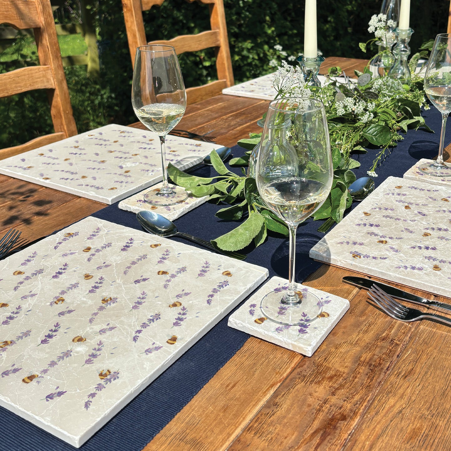 An al fresco dining table set with large marble placemats and coasters featuring a watercolour pattern of busy bees and lavender flowers.