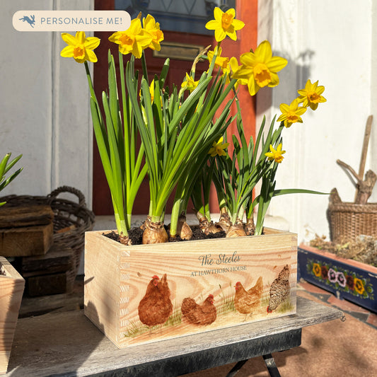 A rectangle wooden planter full of bright daffodils. The planter box shows a watercolour design of a flock of hens, and is personalised with a custom name and house name or number. A perfect gift for a housewarming or new home. Ideal as a first home gift.