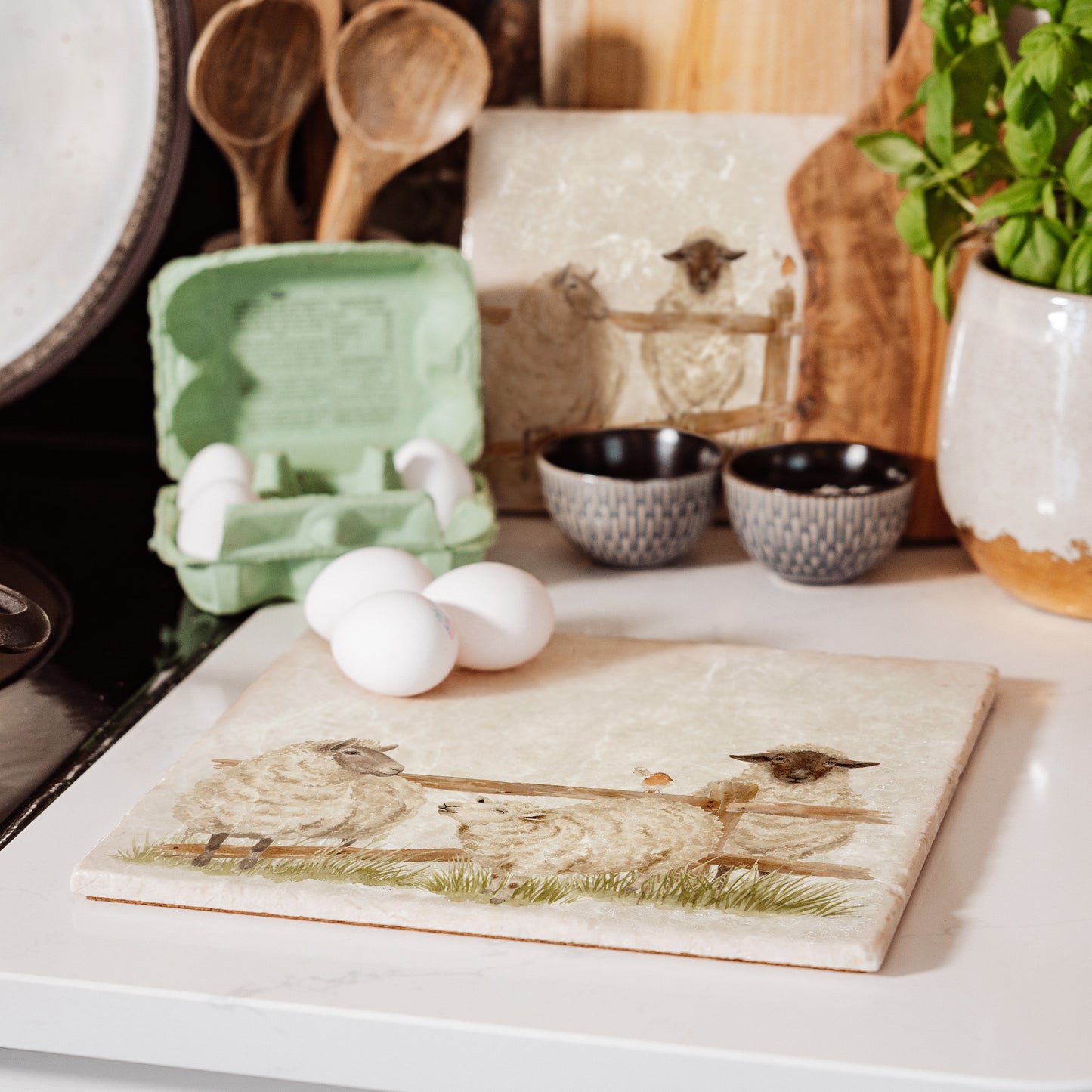 A large marble worktop saver featuring a watercolour design of three sheep escaping from their field through a broken fence. The platter is set on a kitchen work surface next to the hob.
