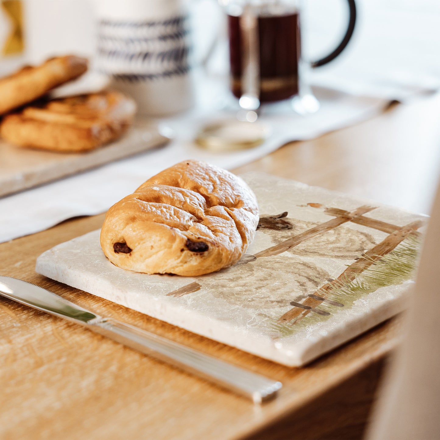 A medium marble platter featuring a watercolour design of two sheep escaping from their field. The platter is set on a country dining table to serve pastries.