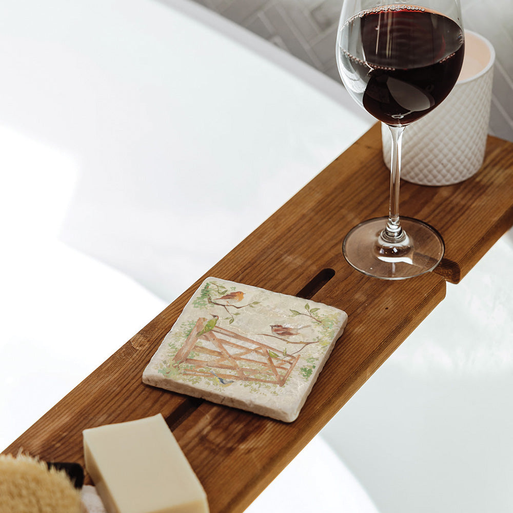 A square marble coaster on a wooden bath tidy above a bubble bath. The coaster features a watercolour design of garden birds in the hedgerow around a paddock gate.